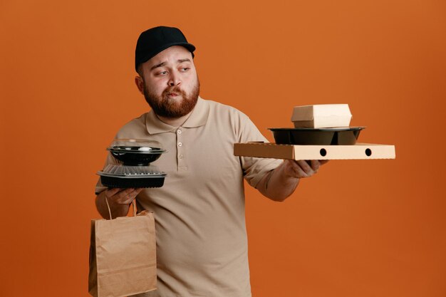 Delivery man employee in black cap and blank tshirt uniform holding food containers with paper bag looking confused having doubts standing over orange background