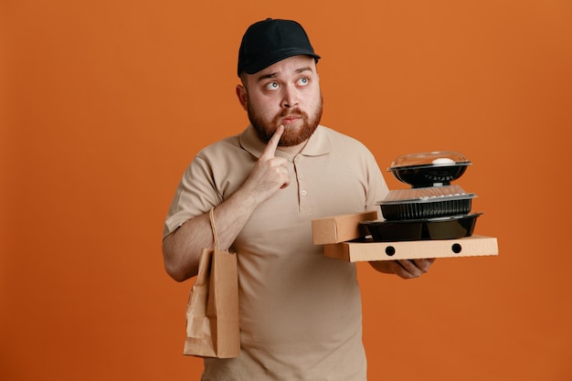 Delivery man employee in black cap and blank tshirt uniform holding food containers with paper bag looking up puzzled thinking standing over orange background