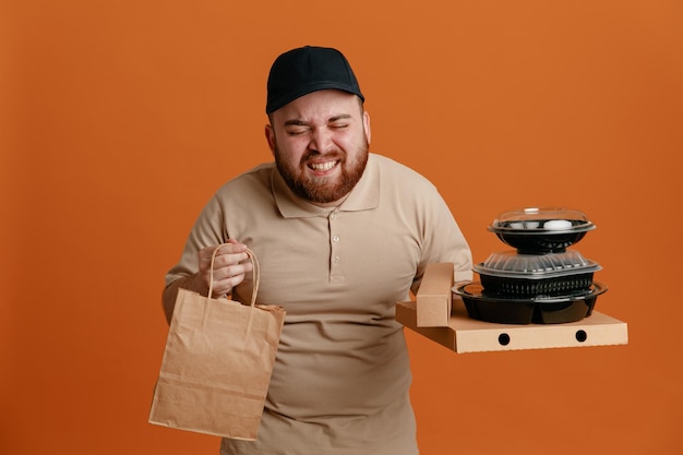 オレンジ色の背景の上に立ってイライラしているように見える紙袋と食品容器を保持している黒い帽子と空白のtシャツの制服を着た配達人の従業員