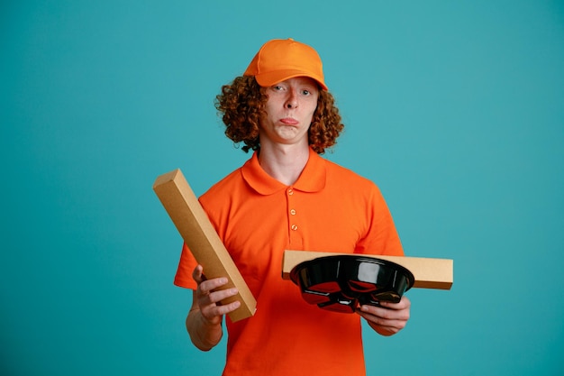Сотрудник доставщика в оранжевой кепке с пустой футболкой держит коробку с пиццей и контейнеры с едой, глядя в камеру, смущенный грустным выражением лица, стоящим на синем фоне
