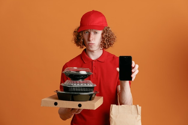 사진 빨간 모자 빈 티셔츠를 입은 배달원 직원이 피자 상자 음식 용기를 들고 오렌지색 배경 위에 혼란스러워 보이는 스마트폰을 보여주는 종이 봉지를 들고 있습니다.