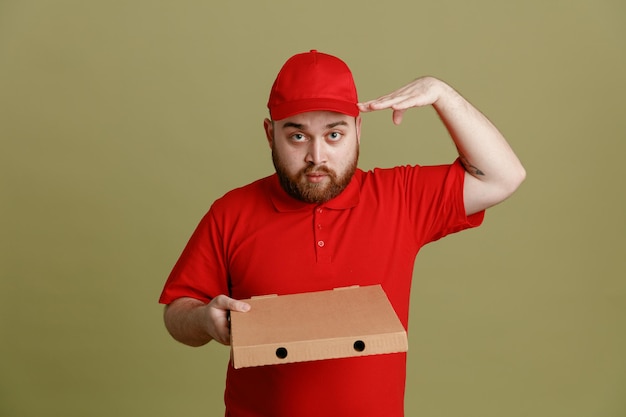 Сотрудник службы доставки в красной кепке с пустой футболкой держит коробку с пиццей и смотрит в камеру с серьезным уверенным выражением лица, держась за руку возле головы и салютуя стоя на зеленом фоне
