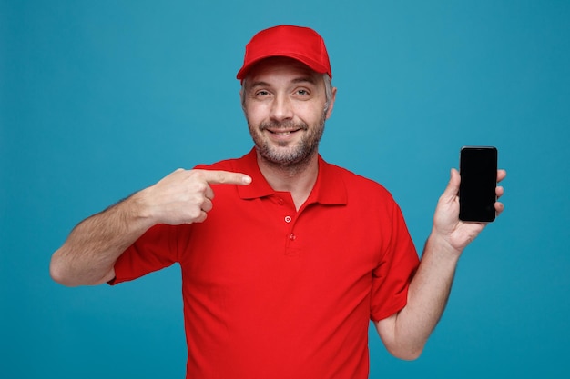 写真 青い背景の上に立っている笑顔のカメラを見て人差し指でそれを指しているスマートフォンを保持している赤い帽子の空白の t シャツの制服を着た配達員従業員