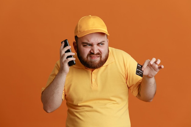 Сотрудник доставщика в желтой кепке с пустой футболкой держит кредитную карту, разговаривает по мобильному телефону, злой и расстроенный, стоя на оранжевом фоне