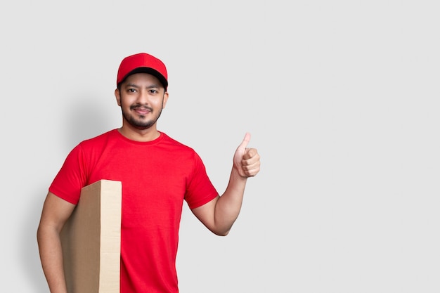 Delivery man employee in red cap blank t-shirt thumbsup uniform hold empty cardboard box isolated on white background