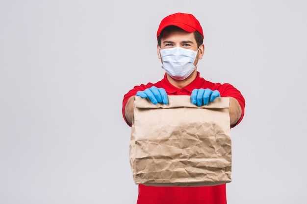 Delivery man employee in red cap blank t-shirt uniform face mask gloves hold empty cardboard box isolated on white wall. Service quarantine pandemic coronavirus virus 2019-ncov concept.