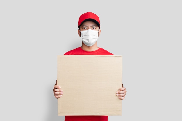 Delivery man employee in red cap blank t-shirt uniform face mask hold empty cardboard box isolated on white background