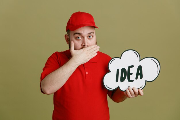 Delivery man employee in red cap blank tshirt uniform holding bubble speech with word idea looking at camera amazed and surprised covering mouth with hand standing over green background