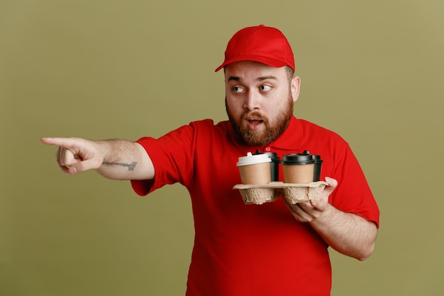 Delivery man employee in red cap blank tshirt uniform holding coffee cups looking aside pointing with index finger at something being surprised standing over green background