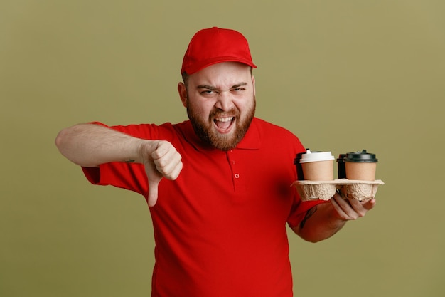 Delivery man employee in red cap blank tshirt uniform holding coffee cups looking at camera angry and frustrated showing thumb down standing over green background