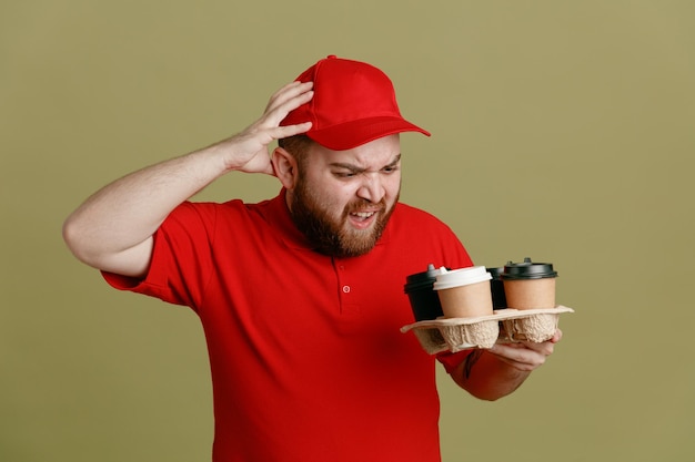 Delivery man employee in red cap blank tshirt uniform holding coffee cups looking at them being confused and annoyed holding hand on his head standing over green background