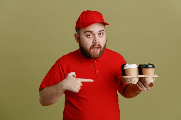 Delivery man employee in red cap blank tshirt uniform holding coffee cups pointing with index finger at cups looking at camera surprised standing over green background