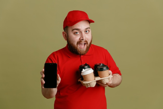 Delivery man employee in red cap blank tshirt uniform holding coffee cups showing smartphone looking at camera happy and positive smiling standing over green background