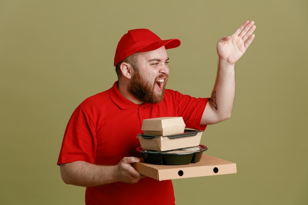 Delivery man employee in red cap blank tshirt uniform holding food containers and pizza box looking aside shouting with aggressive expression raising arm in displeasure standing over green background