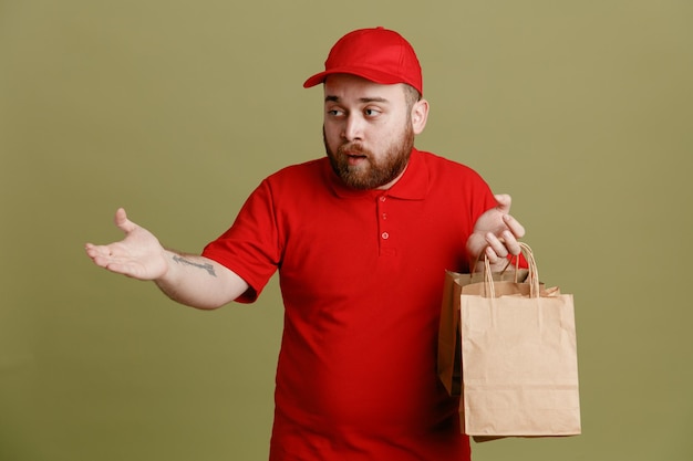 Delivery man employee in red cap blank tshirt uniform holding paper bags looking aside confused raising arm in displeasure standing over green background