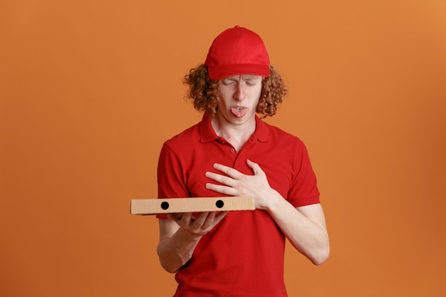 Delivery man employee in red cap blank tshirt uniform holding pizza box looking at it with disgusted expression sticking out tongue standing over orange background