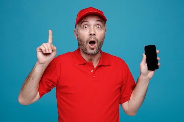 Dipendente dell'uomo delle consegne in uniforme della maglietta vuota con cappuccio rosso che tiene lo smartphone puntato con il dito indice in alto guardando la fotocamera stupito e sorpreso in piedi su sfondo blu