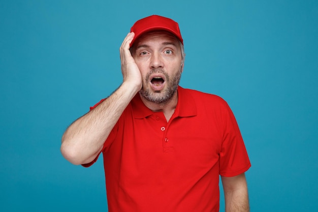Delivery man employee in red cap blank tshirt uniform looking at camera amazed and surprised holding hand on his cheek standing over blue background