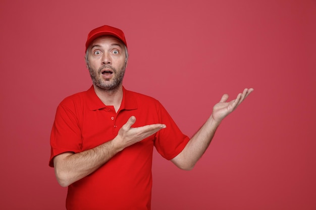 Delivery man employee in red cap blank tshirt uniform looking at camera amazed and surprised presenting with arms of his hands standing over red background