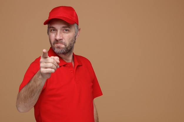 Delivery man employee in red cap blank tshirt uniform looking at camera happy and positive pointing with index finger at camera smiling standing over brown background