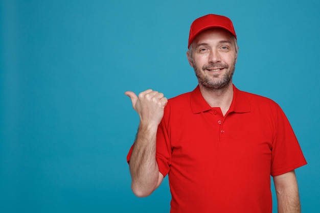 Delivery man employee in red cap blank tshirt uniform looking at camera happy and positive pointing with thumb to the side smiling standing over blue background