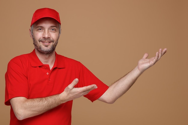 Delivery man employee in red cap blank tshirt uniform looking at camera happy and positive smiling cheerfully presenting with arms of his hands something standing over brown background