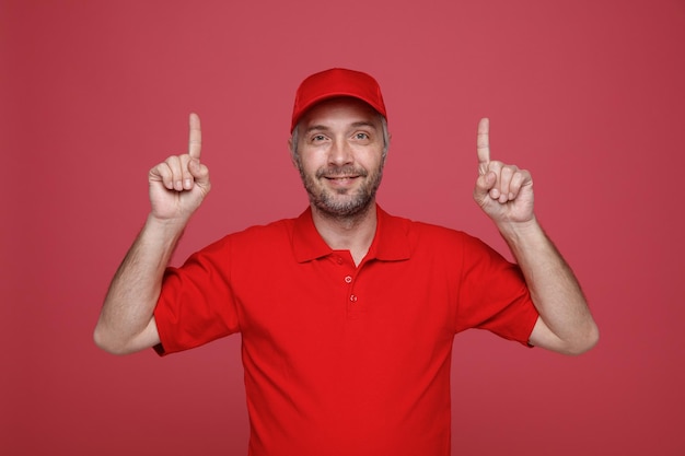 Delivery man employee in red cap blank tshirt uniform looking at camera happy and positive smiling friendly pointing with index fingers up standing over red background