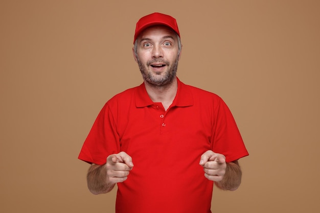 赤い帽子の空白のTシャツの制服を着た配達人の従業員は、茶色の背景の上に立っているカメラに両手で人差し指で指している幸せで前向きな笑顔のカメラを見て