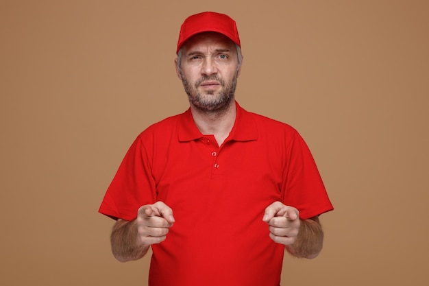 Delivery man employee in red cap blank tshirt uniform looking at camera offended frowning pointing with index fingers with both hands at camera standing over brown background