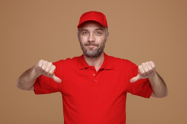 Uomo di consegna dipendente in berretto rosso maglietta vuota uniforme guardando la fotocamera sorridente soddisfatto di sé che punta a se stesso in piedi su sfondo marrone