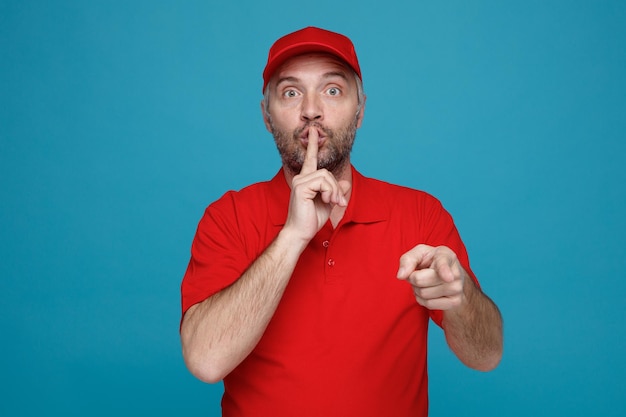 Сотрудник доставщика в красной кепке с пустой футболкой смотрит в камеру, удивленно делая жест молчания, прижимая палец к губам, указывая указательным пальцем на камеру, стоящую на синем фоне