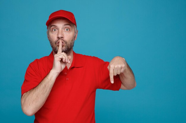 Delivery man employee in red cap blank tshirt uniform looking at camera surprised making silence gesture with finger on lips pointing with index finger down standing over blue background