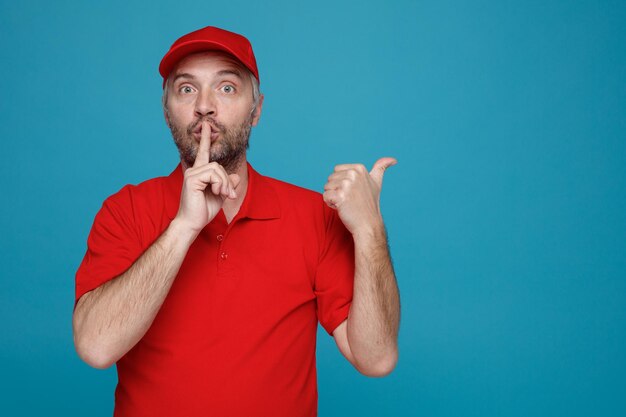 Delivery man employee in red cap blank tshirt uniform looking at camera surprised making silence gesture with finger on lips pointing with thumb to the side standing over blue background