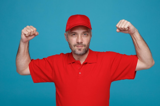 Delivery man employee in red cap blank tshirt uniform looking at camera with confident expression raising clenched fists standing over blue background