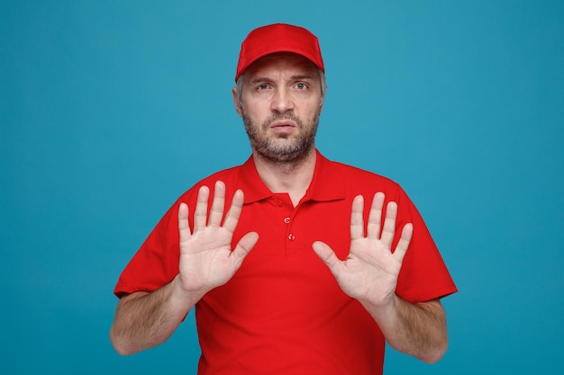 빨간 모자를 쓴 빈 티셔츠를 입은 배달원 직원은 파란 배경 위에 손을 놓고 심각한 얼굴로 정지 제스처를 취하는 카메라를 쳐다보고 있다