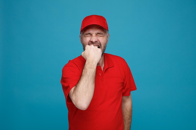 빨간 모자 빈 티셔츠를 입은 배달원 직원은 파란색 배경 위에 서 있는 그의 주먹을 물고 스트레스를 받고 긴장해 보인다