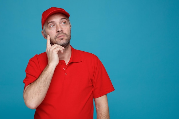 Dipendente dell'uomo delle consegne in uniforme della maglietta vuota con cappuccio rosso che osserva in su con l'espressione pensierosa pensando in piedi su sfondo blu