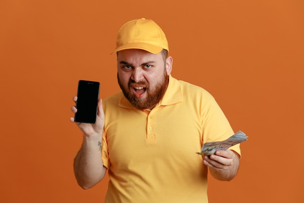 Delivery man employee in yellow cap blank tshirt uniform holding cash and smartphone looking at camera with angry face standing over orange background