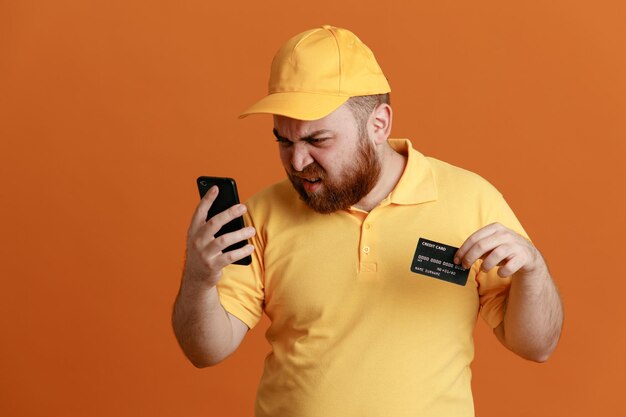 Dipendente dell'uomo di consegna in uniforme della maglietta vuota con cappuccio giallo che tiene la carta di credito guardando il telefono cellulare arrabbiato e deluso in piedi su sfondo arancione