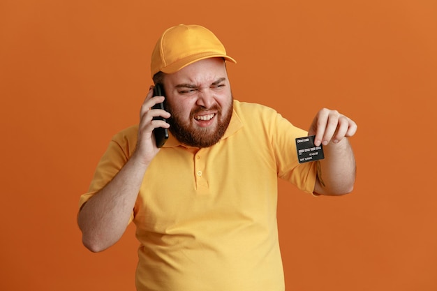 Delivery man employee in yellow cap blank tshirt uniform holding credit card talking on mobile phone angry and annoyed standing over orange background