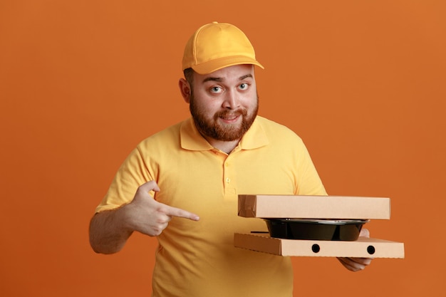 Delivery man employee in yellow cap blank tshirt uniform holding food container and pizza boxes pointing with index finger at boxes smiling confident standing over orange background