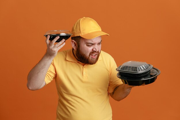 オレンジ色の背景の上に立ってイライラして怒っているように見える食品容器を保持している黄色のキャップの空白のTシャツの制服を着た配達人の従業員