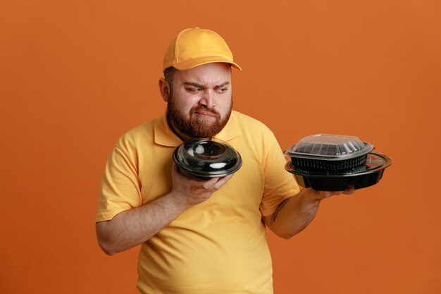 Delivery man employee in yellow cap blank tshirt uniform holding food containers looking confused trying to make choice standing over orange background