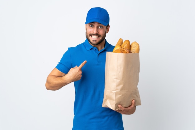Доставщик, держащий сумку, полную хлеба, изолированную на белой стене с удивленным выражением лица