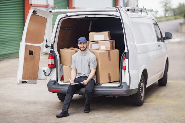 Photo delivery man holding clipboard while sitting in the cargo area of his van