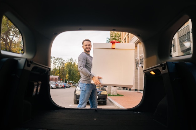 Delivery man holding large festive box with red bow near car outdoors and looking at camera