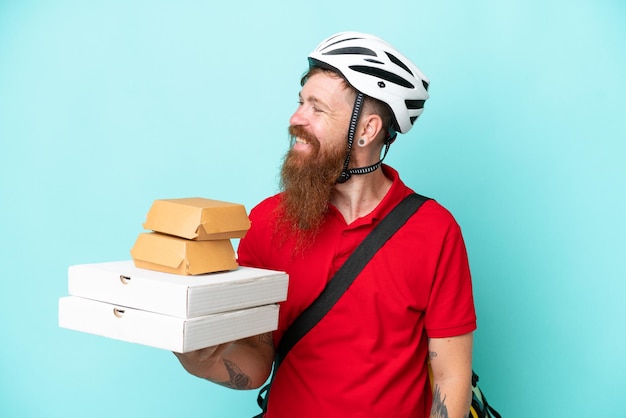 Delivery man holding pizzas and burgers isolated on blue background looking to the side and smiling