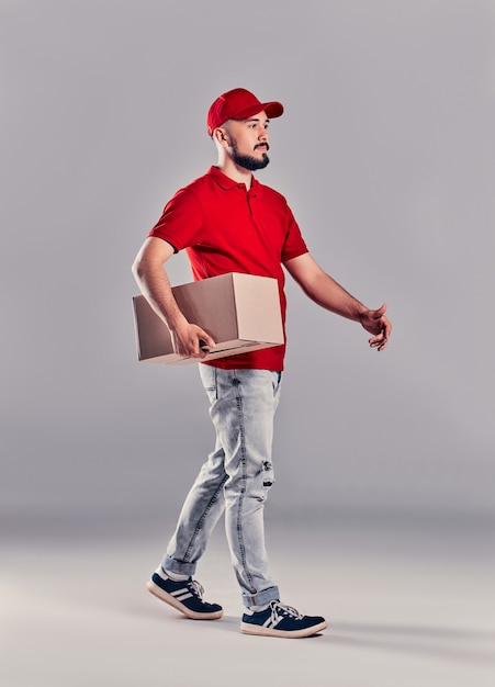 Фото Экспедитор в красной форме, изолированные на сером фоне, студийный портрет. сотрудник-мужчина в принте футболки кепки, работая как дилер-курьер, держит пустую картонную коробку. концепция обслуживания. копировать пространство для копирования