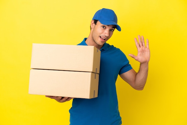 Delivery man over isolated yellow wall saluting with hand with happy expression