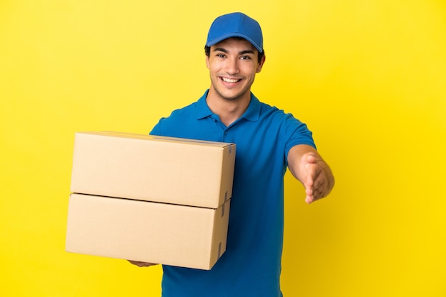 Delivery man over isolated yellow wall shaking hands for closing a good deal